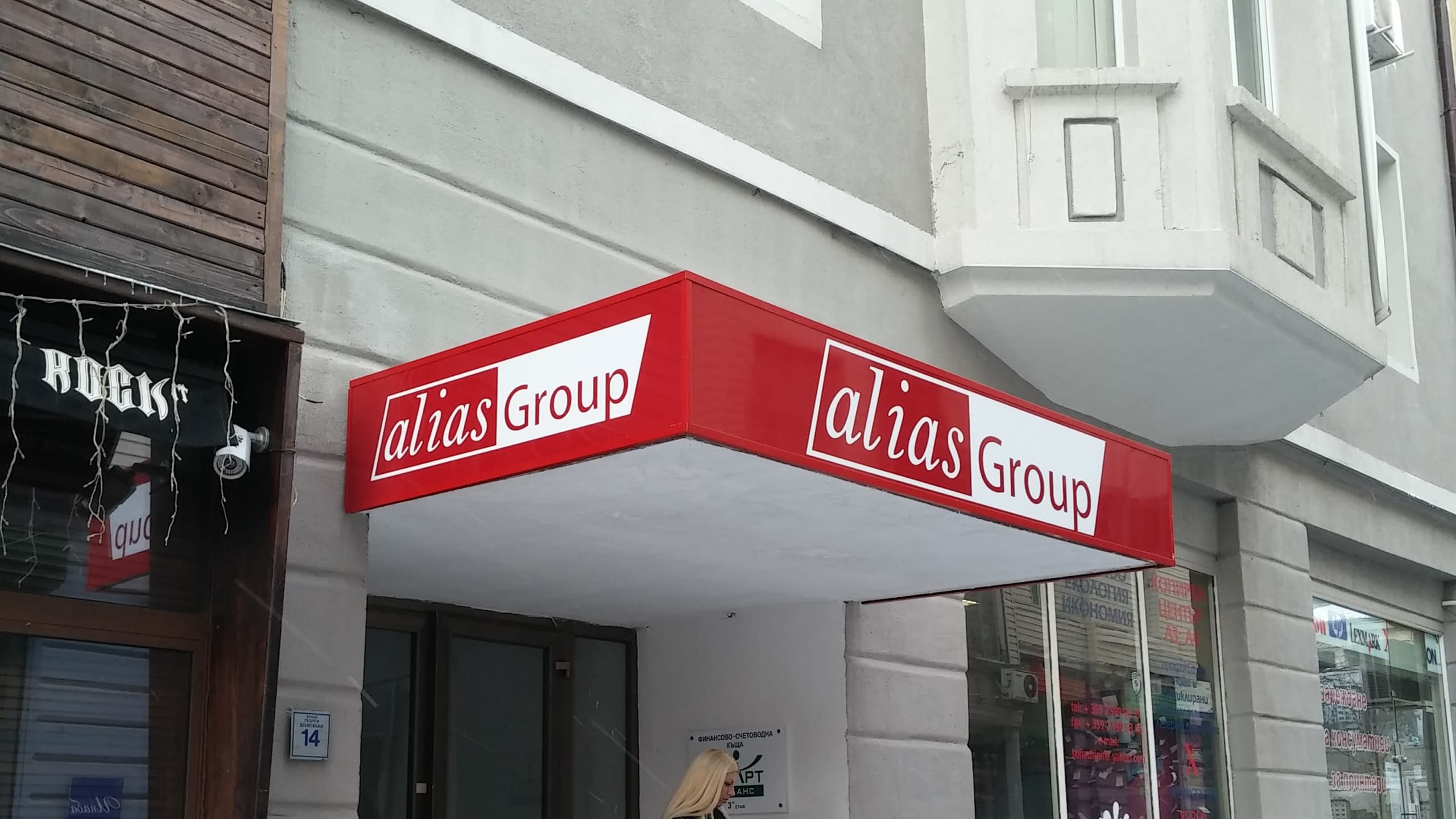 Обемна светеща рекламна табела тип козирка за "alias Group". Червена правоъгълна табела с бяло лого, монтирана над вход на офис или магазин.