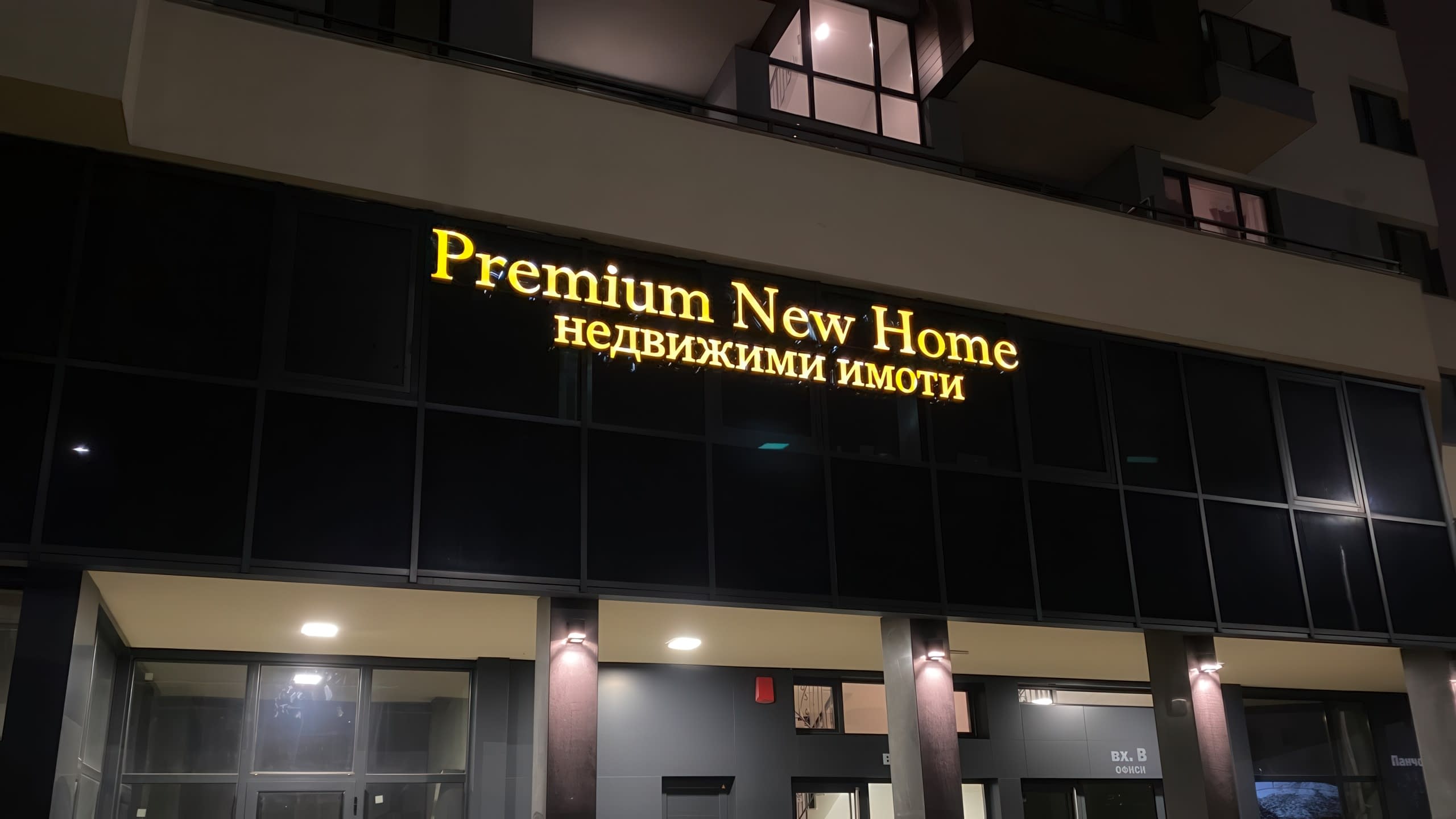 Светеща реклама със златни букви за агенция за недвижими имоти "Premium New Home", монтирана на тъмна фасада на сграда.